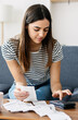 © Xavier Lorenzo - Vertical shot of young adult woman calculating household tax bills and expenses sitting on sofa at home