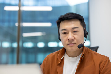 Canvas Print - Asian man businessman wearing headset engaged in virtual meeting. Office environment, focus on laptop screen, slight smile while listening. Represents communication, technology, productivity.