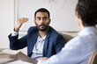 © fizkes - Inspired young Indian business man talking to male coworker in office boardroom, consulting financial advisor, sharing ideas for brainstorming, speaking at table with emotional hands moving