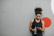 © Prostock-studio - Music and sport. Smiling muscular african american young woman with headphones, smart watch and bottle of water looks at smartphone on wall background, free space