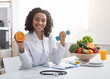 © Prostock-studio - Sport and vitamins for immunity. Happy black woman doctor holding orange and barbell in clinic, posing at her workplace, free space