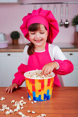 Funny happy little beautiful girl in a chef costume eating popcorn while sitting at the dinner table in the kitchen.	