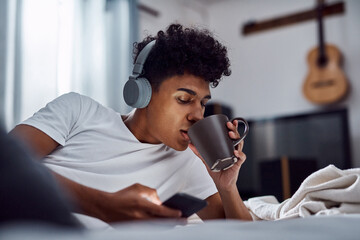 Drinking coffee, phone and man with headphones for music, morning or scroll on social media to relax. Mobile, tea and person in bed for streaming sound, text message or listen to rock in bedroom home