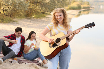 Wall Mural - Young woman playing guitar with her friends on beach at barbeque party