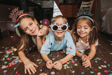 Three small children celebrate their birthday at home, lying on the wooden floor with confetti