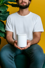 Young man using a portable ai powered language translator while sitting on a pouf, smiling against a bright yellow background with a green plant