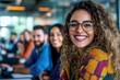 © DKPhoto - A cheerful woman with curly hair and stylish glasses smiles in a lively office environment, surrounded by colleagues, reflecting positivity and teamwork.