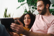 © Studio Marmellata - A couple sitting together on a couch, looking at a tablet in a bright and modern living room filled with greenery, enjoying a cozy and relaxed moment at home.