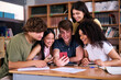 © CarlosBarquero - Diverse group of high school young students looking smiling at mobile phone together in library. Cheerful Generation Z classmates multiracial friends sitting with red cell taking break from class work