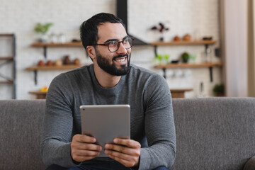 Happy Arabian middle aged man using digital tablet relaxing on couch at home. Adult male user holding tab computer holding pad technology device sitting on sofa in living room looking away. Copy space