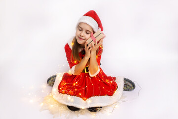 A joyful girl in a festive red dress and Santa hat embraces a gift surrounded by twinkling lights during the holiday season