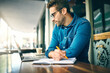 © R Jordaan/peopleimages.com - Laptop, writing and business man in cafe for planning, online research and project ideas. Professional, remote work and person on computer for internet, website and proposal with notes in coffee shop