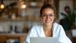 © Lens Legacy - A woman with glasses in a white robe uses a laptop in a cozy setting, suggesting a productive day in comfort, with elements of living space visible around her.