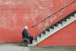 © youriy - An elderly man in a wheelchair beside a red wall, facing a concrete staircase, symbolizing physical barriers and accessibility issues
