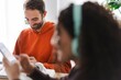 © Studio Marmellata - A man in an orange hoodie smiles down as he writes in a notebook with a woman out of focus in the foreground wearing headphones, adding to the relaxed environment.