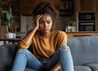 ©     L A S T S H O T - A young woman sits on her sofa with a distressed expression in a cozy living room setting wearing casual clothing Her hand rests on her head as she contemplates with worry and tension