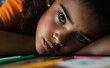 © Curioso.Photography - A young girl, looking tired and thoughtful, rests her head on a desk surrounded by school supplies.