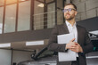 © anatoliycherkas - Modern bearded man in glasses and suit vehicle sales consultant using laptop inside car dealership