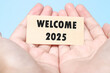 © Scorpeow - Hand holding a note Welcome 2025 with a blue background. New year concept with typographic 2025. Motivational words to welcome the new year 2025