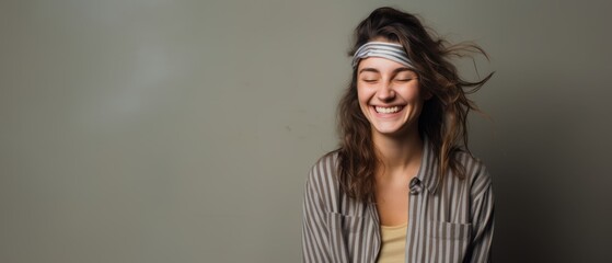 Wall Mural - A delighted woman closes her eyes, smiling joyfully while wearing a headband. Her carefree expression radiates positivity against a simple grey backdrop.