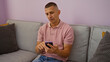 © Krakenimages.com - Young man using smartphone seated on a gray couch in a cozy, tidy living room with purple walls, wearing a pink polo shirt and jeans, focused on his screen with a thoughtful expression.