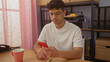 © Krakenimages.com - Young hispanic man in office checking mobile phone while sitting at desk with a pink coffee mug and documents in the background.