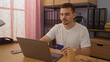 © Krakenimages.com - Young hispanic man with a beard working on a laptop holding a credit card in a modern office with shelves, binders, and coffee cup on a desk.