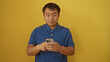© Krakenimages.com - A handsome young chinese man in a blue shirt attentively uses his smartphone against an isolated yellow background.