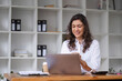 © wichayada - Confident Businesswoman Working on Laptop in Modern Office Environment with Shelves and Documents in Background