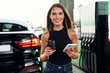 © standret - Fresh cup of coffee in hand, smartphone. A young woman at a gas station with her car