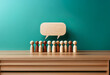 © joeycheung - A row of wooden figures stand in front of a blank speech bubble on a wooden table against a teal wall.
