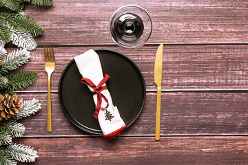 Festive Christmas table setting with black plate, napkin, golden cutlery set and fir tree branches on dark wooden background top view.