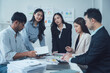 © Kritdanai - Teamwork Makes the Dream Work: A diverse group of young business professionals collaborates around a table, engaged in a serious discussion over documents and charts.