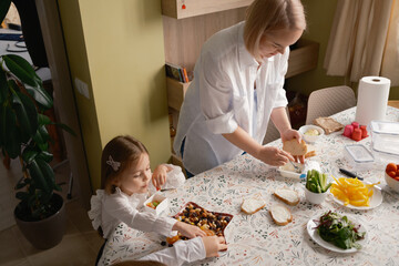 A mother and daughter prepare sandwiches and snacks at the kitchen table, emphasizing healthy, fresh ingredients for school. The scene highlights family togetherness and morning routines.