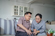 © ultramansk - An elderly couple sits in their living room. The man holds his knee, appearing to be in discomfort, while his partner offers support. The image conveys care, concern, and the bond between the couple.