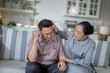 © ultramansk - An elderly couple sits on a couch in a cozy living room. The man holds his head in discomfort, while his partner offers support. The image highlights care, concern, and the bond between the couple.