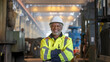 © Tom - A senior Hispanic male construction worker in a bright yellow safety jacket and hard hat smiles confidently, standing inside a large industrial facility.