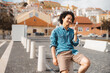 © skyNext - A young man with curly black hair, wearing a denim shirt and brown shorts, sits outdoors on a concrete post, smiling at his smartphone; urban setting with traditional Portuguese buildings behind him