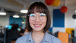 © Tom - Young Asian woman with glasses, smiling warmly in a modern office environment with colorful decor.