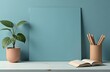 © Yana Gavriloski - A serene workspace displays a green plant in a pot, a container filled with pencils, and an open book resting on a white wooden table, all against a soft blue wall