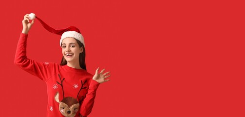 Wall Mural - Young woman in Christmas sweater and with Santa hat on red background with space for text