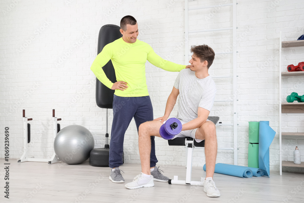 Sporty young man training with dumbbell and coach in gym
