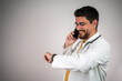 © Nenad - Young hispanic man wearing doctor uniform looking watch holding report at clinic.