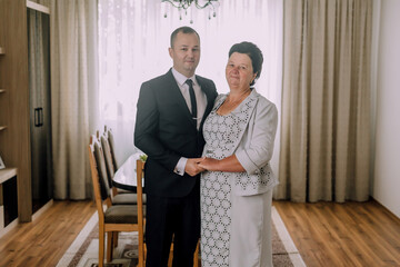 A man and a woman are standing in a room with a dining table in the background. The man is wearing a suit and tie, while the woman is wearing a dress. They are both smiling