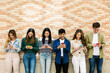 © Xavier Lorenzo - Group of friends using smartphones, standing against a brick wall, enjoying social media and mobile technology. Youth and technology lifestyle concept