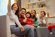 © Garun Studios - relationship, parenthood, happiness, family, enjoying, happy, care, celebration, christmas, laughing. A family of five is sitting on a couch in a living room, smiling and waving at the camera.