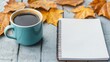 © Bantita - A cozy autumn scene featuring a cup of coffee beside a blank notepad and scattered orange leaves on a wooden surface.