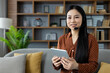 © Liubomir - Asian woman with headset using tablet at home, smiling, seated on couch. Concept of remote work, online communication, technology. Ideal for virtual meeting, customer service, or e-learning.