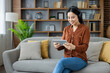 © Liubomir - Asian woman with headphones happily engaged on tablet. Seated on sofa in cozy setting. Relaxed, modern home environment with natural elements. Technology use in comfortable space.