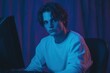 © Sohaib q - A young man with tousled hair sits in front of a computer, illuminated by blue and purple lighting. He has a serious expression, wearing a white long-sleeve shirt, creating a moody atmosphere
